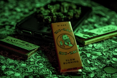 A packet of Zig-Zag branded rolling papers with a vintage illustration on the front is surrounded by other similar products on a textured surface. The lighting casts a greenish hue over the scene, creating a moody atmosphere.