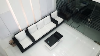 A modern, minimalist waiting area featuring a black and white sofa set arranged along the wall. A sleek black coffee table sits in front of the sofa. Glass office walls are visible on the right side, providing a view into an adjacent room.