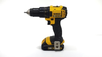 A cordless power drill with a striking yellow and black color scheme. It features a robust build with a metal drill chuck and a visible brand logo. The drill appears to be battery-operated, as indicated by the battery pack at the base.