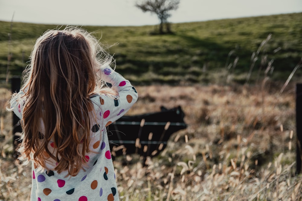 girl wearing white and multicolored polka-dot long-sleeved shirt standing while facing back on green field during daytime