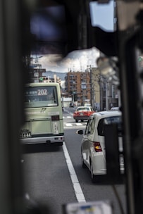 In a busy urban area, vehicles including a bus and cars are moving through a street with buildings and traffic lights. The scene shows the hustle of city life with infrastructure and transport prominently displayed.