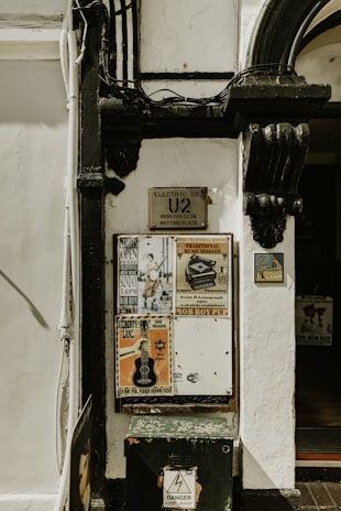 A wall with various posters and signs attached to it. The posters advertise musical events and have vintage designs. There is a sign indicating a meeting place for an Irish fan club, and several signs with text and images. The wall also has a small warning sign at the bottom with the text 'Danger Keep Away'.