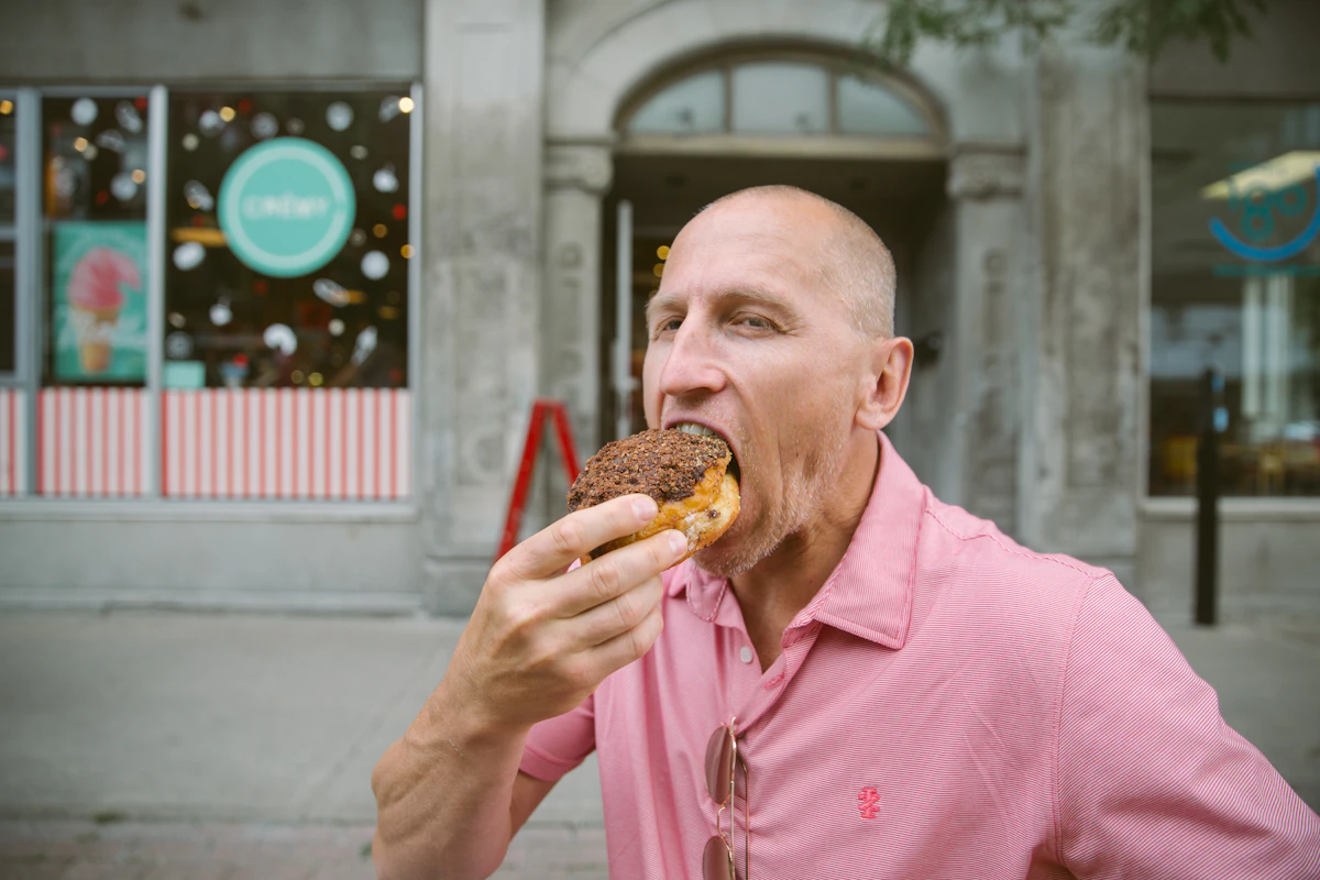 Middle-aged man taking a bite of a sugary chocolate donut on a city street, illustrating how sugar intake affects testosterone production.