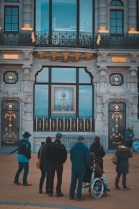 A group of people is gathered outside a grand architectural building with ornate details, including gold accents on a stone exterior. Large windows reflect some interior elements, and a significant framed artwork or city view is visible through the central window. One individual is in a wheelchair, highlighting inclusivity among the crowd.