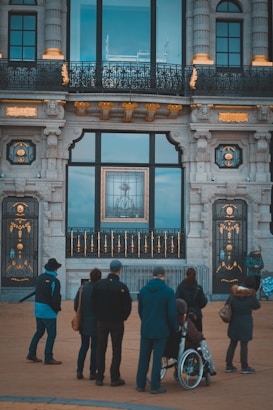 A group of people is gathered outside a grand architectural building with ornate details, including gold accents on a stone exterior. Large windows reflect some interior elements, and a significant framed artwork or city view is visible through the central window. One individual is in a wheelchair, highlighting inclusivity among the crowd.