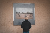 A person wearing a black jacket and beige cap is standing in front of a large photograph displayed on a textured wall. The photograph depicts a cemetery with numerous graves and headstones, set under a cloudy sky. Trees are visible in the background of the photo within the image.