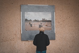 A person wearing a black jacket and beige cap is standing in front of a large photograph displayed on a textured wall. The photograph depicts a cemetery with numerous graves and headstones, set under a cloudy sky. Trees are visible in the background of the photo within the image.