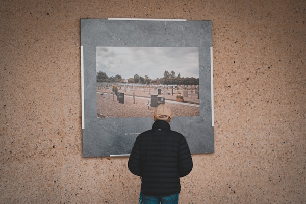 A person wearing a black jacket and beige cap is standing in front of a large photograph displayed on a textured wall. The photograph depicts a cemetery with numerous graves and headstones, set under a cloudy sky. Trees are visible in the background of the photo within the image.