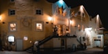 A nighttime scene of a historic building with the signage 'Hotel de la Suc' and 'A.D. 1824'. Warm lights illuminate the stone facade, highlighting flags above the entrance and potted plants by the windows. A small staircase leads up to a terrace where two people stand. The atmosphere suggests a cozy, inviting ambiance of a quaint European hotel or restaurant.