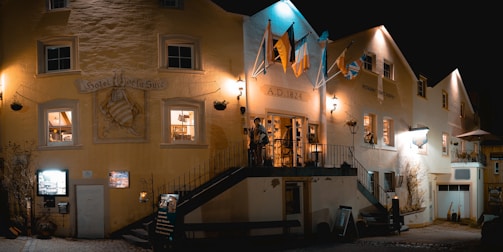 A nighttime scene of a historic building with the signage 'Hotel de la Suc' and 'A.D. 1824'. Warm lights illuminate the stone facade, highlighting flags above the entrance and potted plants by the windows. A small staircase leads up to a terrace where two people stand. The atmosphere suggests a cozy, inviting ambiance of a quaint European hotel or restaurant.
