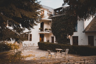 A serene courtyard setting featuring a rustic building with white walls, adorned with green ivy climbing up its facade. A few chairs and a round table are placed on a gravel path, with lush greenery and large trees surrounding the area. The scene conveys a sense of tranquility and seclusion, enhanced by the warm, natural lighting.