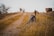 A hiking backpack and trekking poles rest on a grassy path in an open, natural setting. The background features a blurred trail, scattered trees, and an overcast sky.