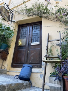 An old wooden door is surrounded by climbing plants and greenery. A sign with contact information is visible near the door. A blue backpack rests on the stone steps leading up to the door. A ladder with potted plants stands beside the entrance.