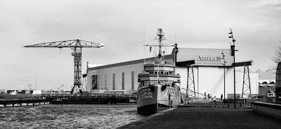 A black and white image of a harbor scene featuring a ship docked alongside an industrial building with the sign 'Amels Yacht Building.' A large crane is visible to the left, extending over the water, and several people can be seen walking on the pier. The water is calm, and the sky appears overcast.