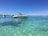 A sleek white speedboat is anchored in crystal clear turquoise waters, with another boat visible in the background. The scene is set against a backdrop of a distant shoreline and a bright blue sky scattered with a few clouds.