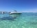 A sleek white speedboat is anchored in crystal clear turquoise waters, with another boat visible in the background. The scene is set against a backdrop of a distant shoreline and a bright blue sky scattered with a few clouds.