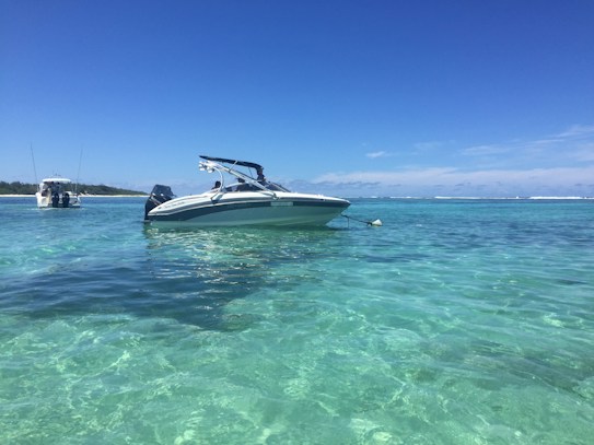 A sleek white speedboat is anchored in crystal clear turquoise waters, with another boat visible in the background. The scene is set against a backdrop of a distant shoreline and a bright blue sky scattered with a few clouds.