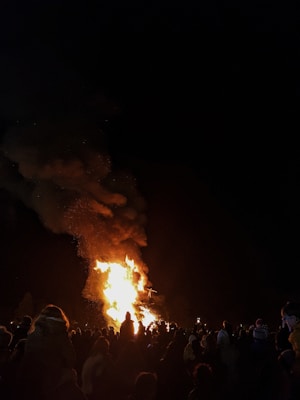 A large crowd gathers outdoors at night, focusing on a towering bonfire emitting bright flames and thick smoke. Silhouettes of people are visible against the backdrop of the fire, creating a dramatic contrast between the darkness and the intense light. Some attendees are capturing the event on their phones.