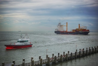 A pilot boat is navigating through a body of water alongside a large cargo ship carrying multiple containers. The pilot boat has a vibrant red and white color scheme, with the word 'PILOT' clearly visible on its side. The cargo ship is significantly larger, with cranes and numerous stacked containers in various colors. The scene is set against an overcast sky, and a dock with posts is visible in the foreground.