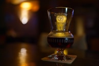 A glass with dark liquid stands on a coaster on a surface with a blurred, dimly lit background. The glass is embossed with text and a logo, suggesting a brand of beverage.