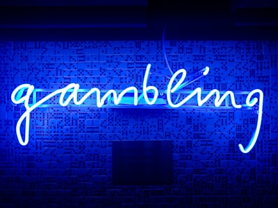 A bright blue neon sign spells out the word 'gambling' in cursive script. The background features a textured wall covered in domino tiles, also illuminated by the blue light, creating a vivid and visually striking scene.