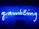 A bright blue neon sign spells out the word 'gambling' in cursive script. The background features a textured wall covered in domino tiles, also illuminated by the blue light, creating a vivid and visually striking scene.