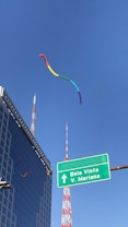 A rainbow-colored kite is flying high against a clear blue sky. In the background, there are radio towers and a modern office building with glass windows. A green road sign points towards 'Bela Vista' and 'V. Mariana'.