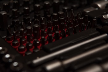 A collection of various dark-colored metal tool bits and attachments arranged closely together on a red background. The lighting creates a contrasting effect, highlighting the shapes and edges of the tools.