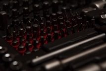A collection of various dark-colored metal tool bits and attachments arranged closely together on a red background. The lighting creates a contrasting effect, highlighting the shapes and edges of the tools.