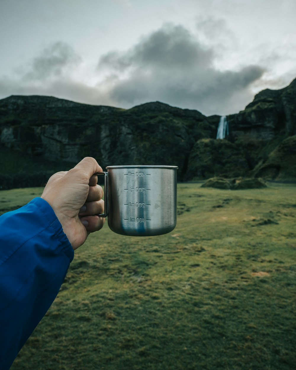 person holding out gray metal cup near waterfalls