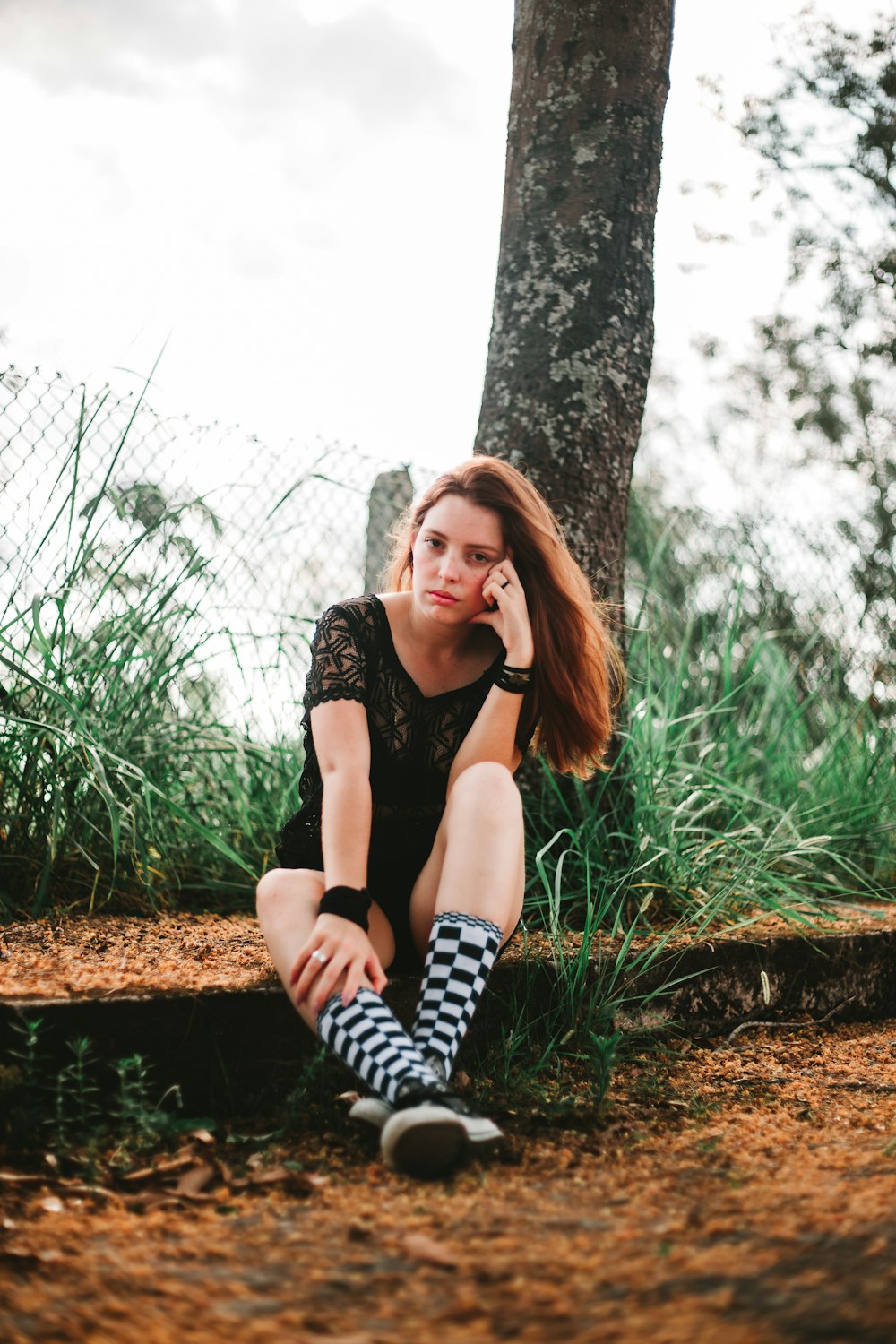 selective focus photography of sitting woman beside tree and chain link fence during daytime