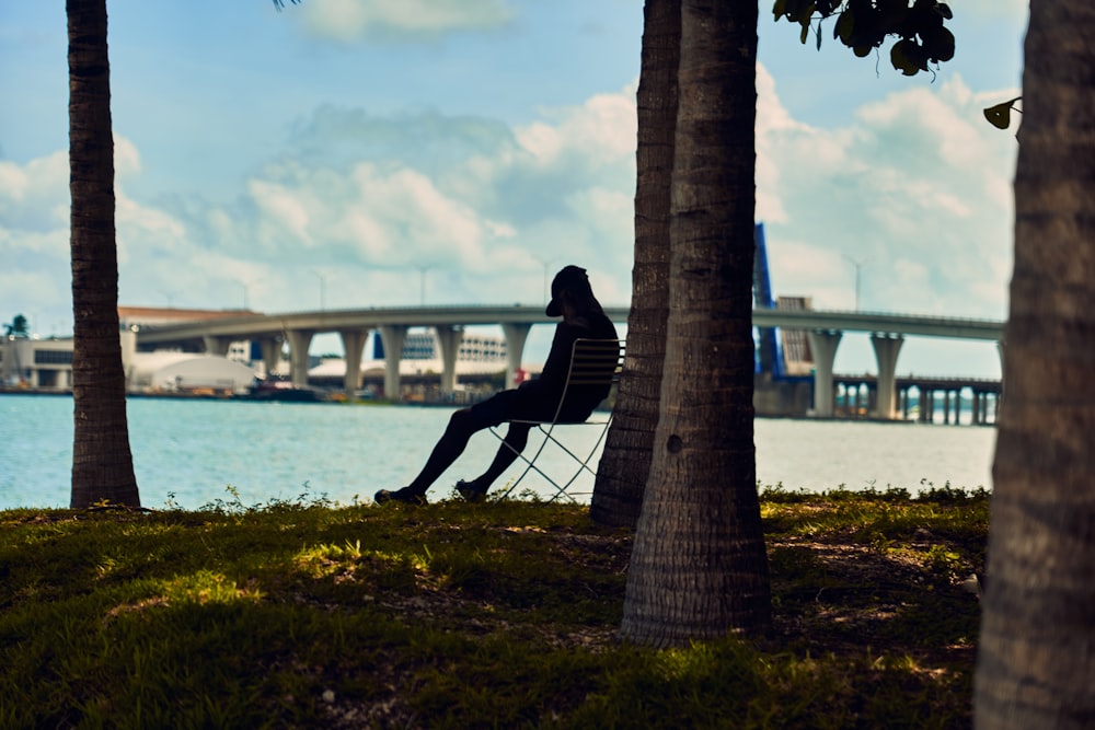 Person mit Mütze sitzt tagsüber auf einem Stuhl unter einem Baum neben einem Gewässer