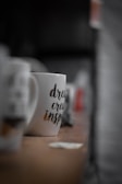 A white mug with printed words 'dream, create, inspire' is placed on a wooden surface. The mug is in focus with a blurred background showing other out-of-focus mugs and objects.