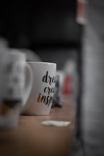 A white mug with printed words 'dream, create, inspire' is placed on a wooden surface. The mug is in focus with a blurred background showing other out-of-focus mugs and objects.