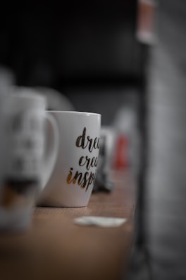 A white mug with printed words 'dream, create, inspire' is placed on a wooden surface. The mug is in focus with a blurred background showing other out-of-focus mugs and objects.
