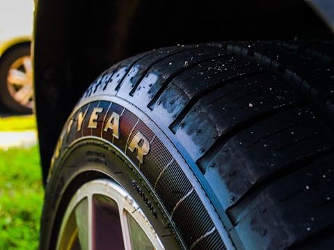 A close-up view of a car tire featuring the brand name in large letters on the side. The tire tread is clearly visible, showing detailed textures and patterns. The background consists of grass and parts of another vehicle, slightly out of focus.
