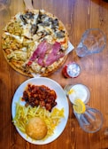 A wooden table is set with a diverse assortment of food and drinks. A pizza covered with a variety of toppings is positioned on a wooden serving board. Nearby, a large plate contains French fries, a bread roll, and a rich stew or casserole with chunks of meat, topped with a savory sauce. A can of soda and two empty glasses, including one glass with a lemon slice and straw, are placed nearby.