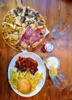 A wooden table is set with a diverse assortment of food and drinks. A pizza covered with a variety of toppings is positioned on a wooden serving board. Nearby, a large plate contains French fries, a bread roll, and a rich stew or casserole with chunks of meat, topped with a savory sauce. A can of soda and two empty glasses, including one glass with a lemon slice and straw, are placed nearby.
