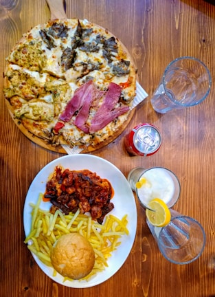 A wooden table is set with a diverse assortment of food and drinks. A pizza covered with a variety of toppings is positioned on a wooden serving board. Nearby, a large plate contains French fries, a bread roll, and a rich stew or casserole with chunks of meat, topped with a savory sauce. A can of soda and two empty glasses, including one glass with a lemon slice and straw, are placed nearby.