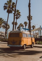 An old, orange camper van is parked in a sunny lot surrounded by tall palm trees. The scene suggests a laid-back, coastal environment with clear blue skies and a calm, relaxed atmosphere.