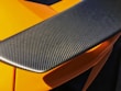 A close-up view of a carbon fiber car spoiler with a textured surface. The orange bodywork of the car can be seen partially underneath, highlighted by sunlight.