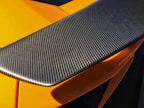 A close-up view of a carbon fiber car spoiler with a textured surface. The orange bodywork of the car can be seen partially underneath, highlighted by sunlight.