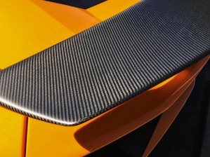 A close-up view of a carbon fiber car spoiler with a textured surface. The orange bodywork of the car can be seen partially underneath, highlighted by sunlight.