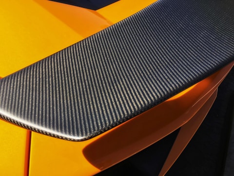 A close-up view of a carbon fiber car spoiler with a textured surface. The orange bodywork of the car can be seen partially underneath, highlighted by sunlight.