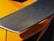 A close-up view of a carbon fiber car spoiler with a textured surface. The orange bodywork of the car can be seen partially underneath, highlighted by sunlight.