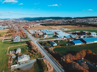 Aerial view of a rural landscape featuring a straight road dividing fields and residential areas. Several houses and agricultural buildings with blue roofs are visible, surrounded by fields and patches of autumn-colored trees. Hills and a distant town can be seen under a clear blue sky with scattered clouds.