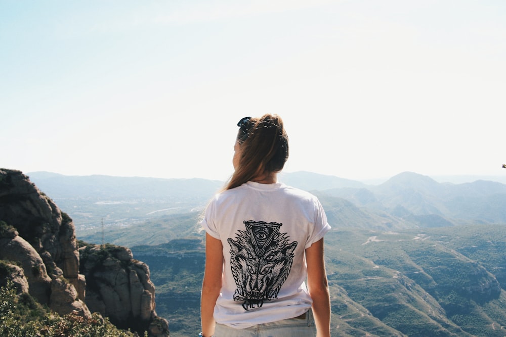 白と黒のTシャツを着た女性が崖に立って昼間、山を眺めている