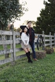 A couple poses by a wooden fence in a rural setting. The woman leans against the fence wearing a white sweater, checked skirt, black knee-high boots, and a black hat on her head. The man stands beside her in a jacket and jeans, his arm resting on the fence. Nearby, a straw hat is perched on the fence post, and a wagon wheel sits next to a bale of hay. The background features trees and a grassy area, suggesting a peaceful countryside atmosphere.