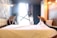 Two people are making a bed in a well-lit modern room. The room features a large window with dark curtains, geometric patterns on one wall, and several hanging light bulbs on the ceiling.