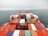 A large number of colorful shipping containers are neatly stacked on a cargo ship that is sailing on an open, calm ocean. The horizon is slightly foggy, giving a sense of distance and vastness.